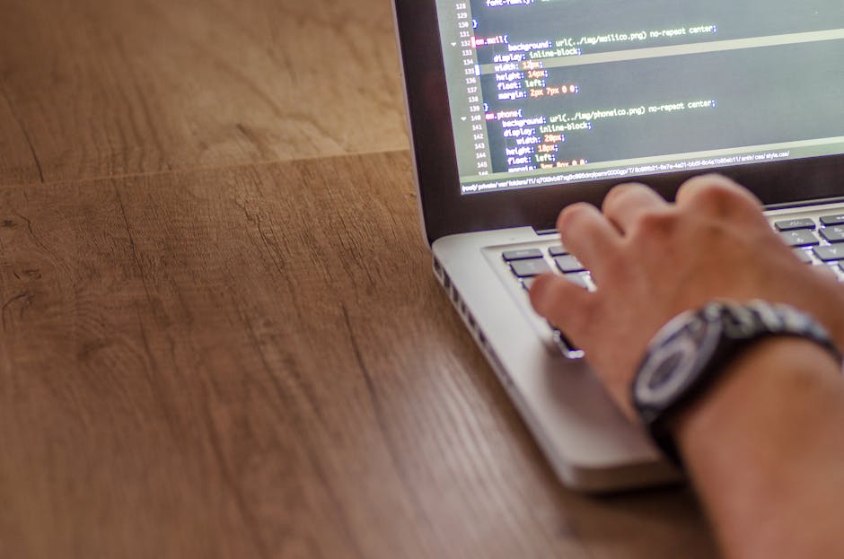 A person's hand with a wristwatch is typing on a laptop keyboard displayed on a wooden surface. The laptop screen shows lines of code or scripting text, indicating activity related to programming or w