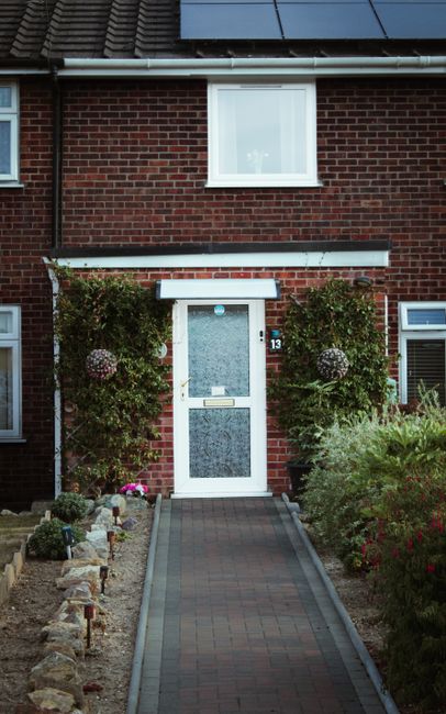 A small front garden in a residential area featuring a paved pathway leading to a white front door with a glass panel, set in a red-brick house. The garden is bordered by a variety of plants, includin